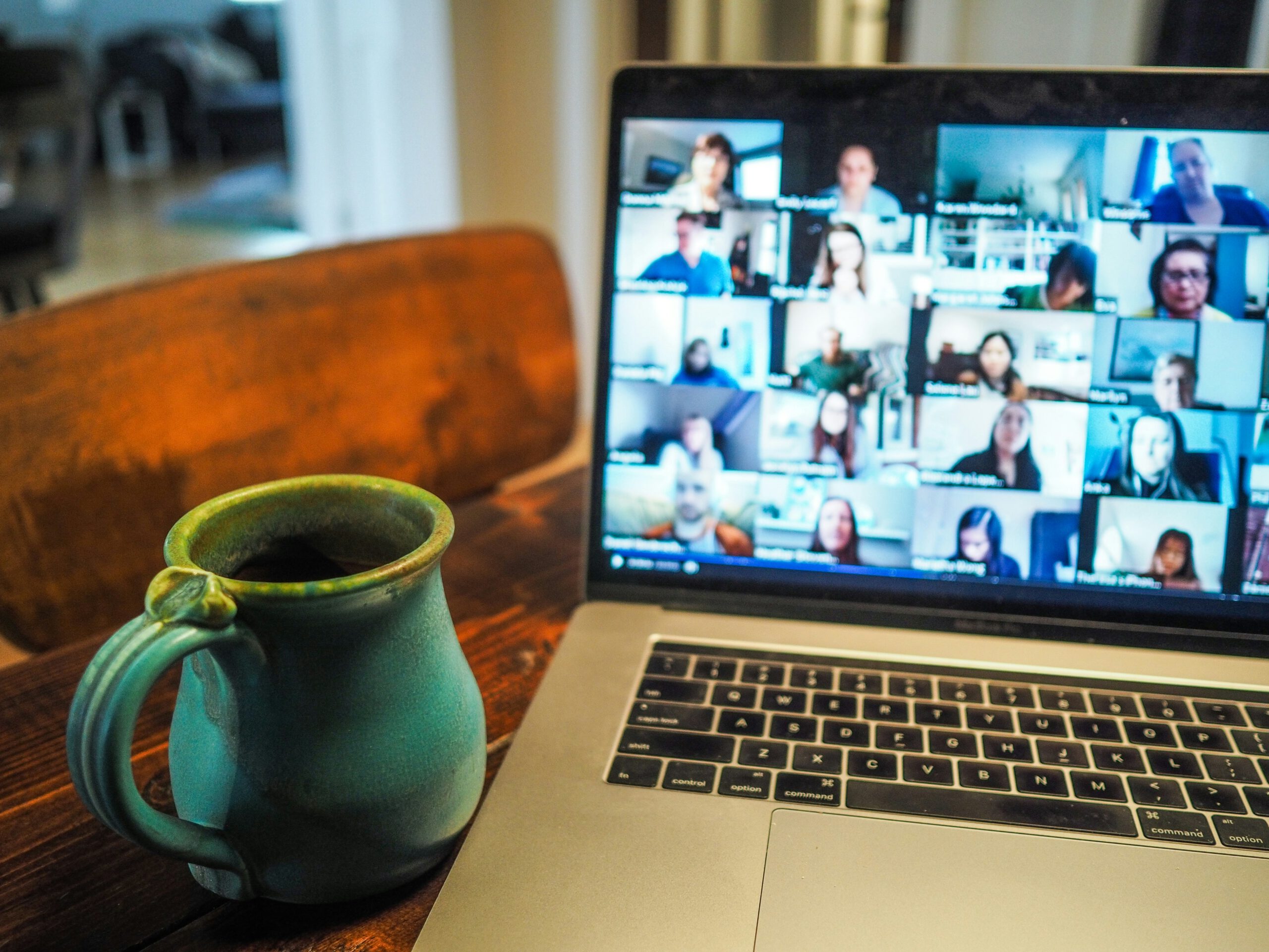 Das Foto zeigt einen Laptop, auf dessen Bildschirm ein Video-Meeting mit vielen Teilnehmern in kleinen Kacheln zu sehen ist. Im Vordergrund steht auf einem Holztisch eine grüne Keramiktasse mit Henkel, die wahrscheinlich Kaffee oder Tee enthält. Der Raum im Hintergrund ist unscharf, was den Fokus auf den Laptop und die Tasse legt. Die Szene vermittelt eine typische Homeoffice- oder Remote-Arbeitssituation.