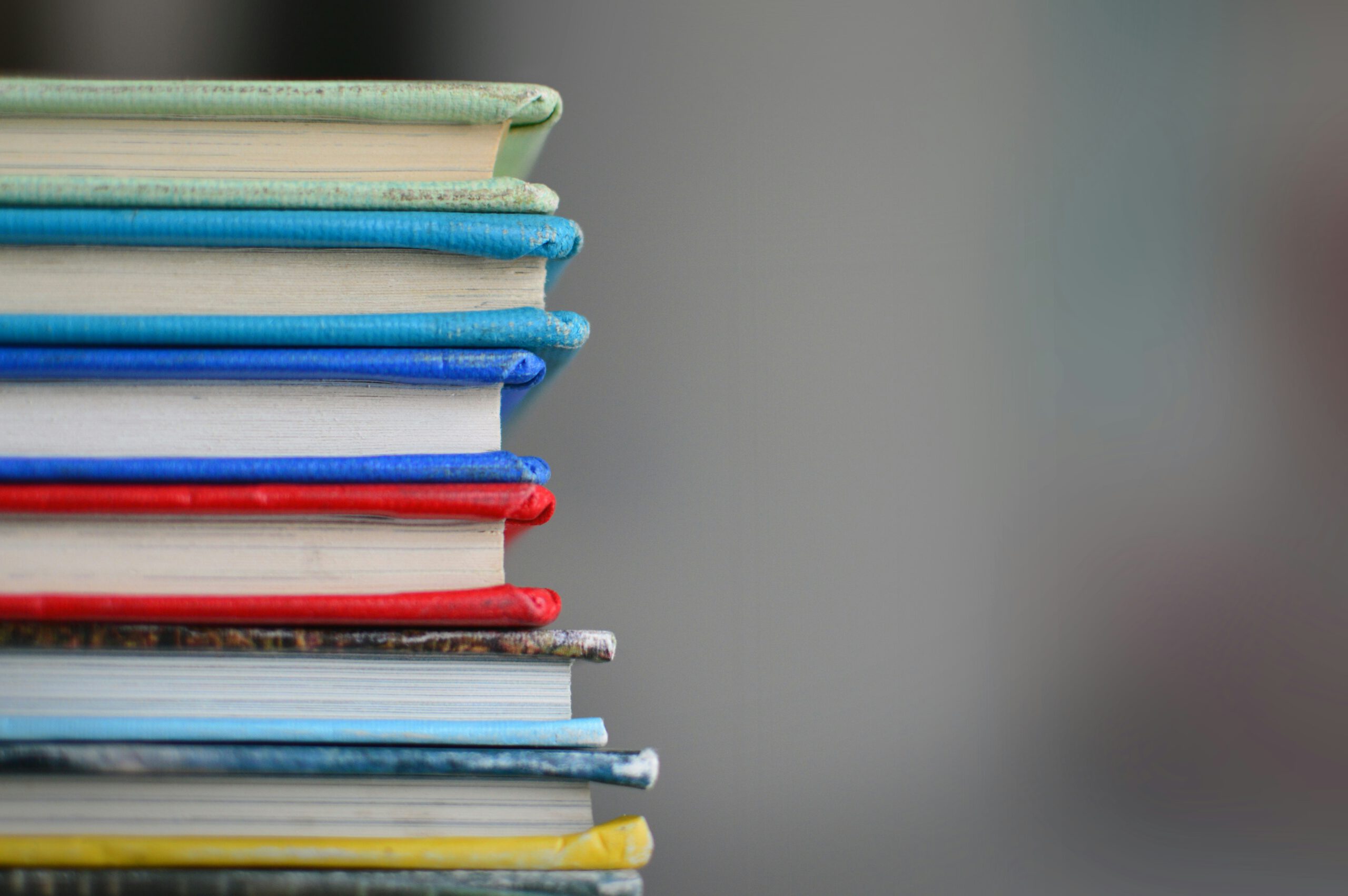 Das Foto zeigt einen Stapel Bücher, die eng aufeinander gestapelt sind. Die Buchrücken sind in verschiedenen Farben gehalten, darunter Blau, Rot und Grün. Die Bücher wirken leicht abgenutzt, was auf häufige Benutzung schließen lässt. Der Hintergrund ist unscharf und lenkt den Fokus auf die Buchrücken, die am linken Bildrand angeordnet sind.