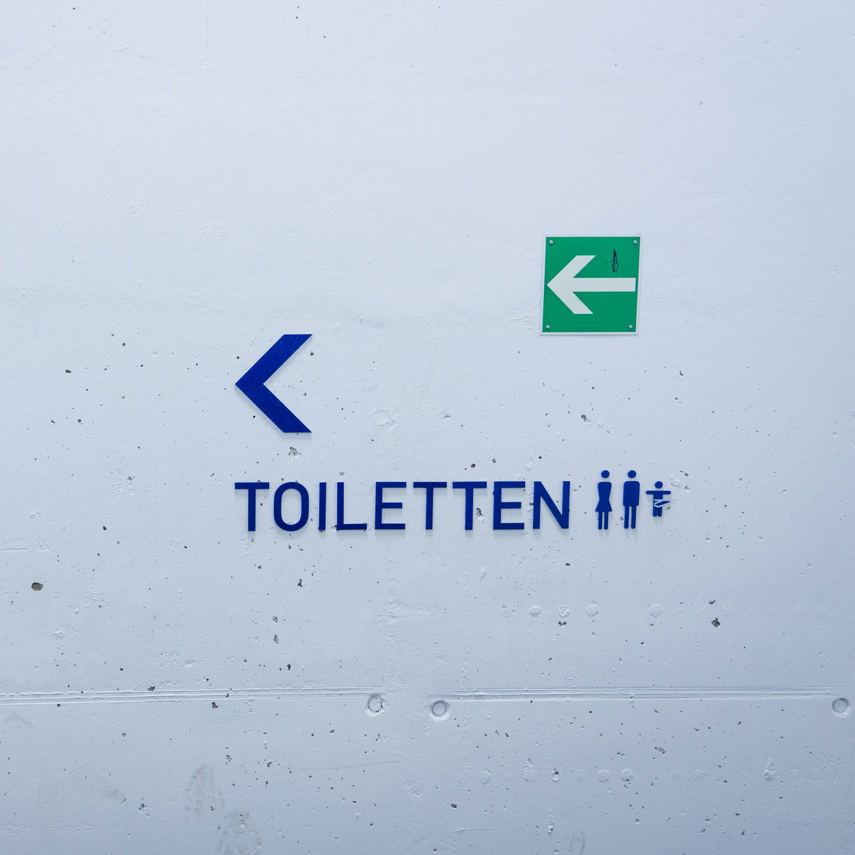 Ein weiß gestrichener Betonwand zeigt ein Schild mit der Aufschrift "Toiletten" in blauen Buchstaben. Links daneben weist ein blauer Pfeil nach links. Rechts oberhalb des Schriftzugs befindet sich ein grünes Schild mit einem weißen Pfeil, das den Fluchtweg in die gleiche Richtung anzeigt. Neben dem Toilettenschriftzug sind Piktogramme für eine Herrentoilette, Damentoilette und eine Wickelstation zu sehen.