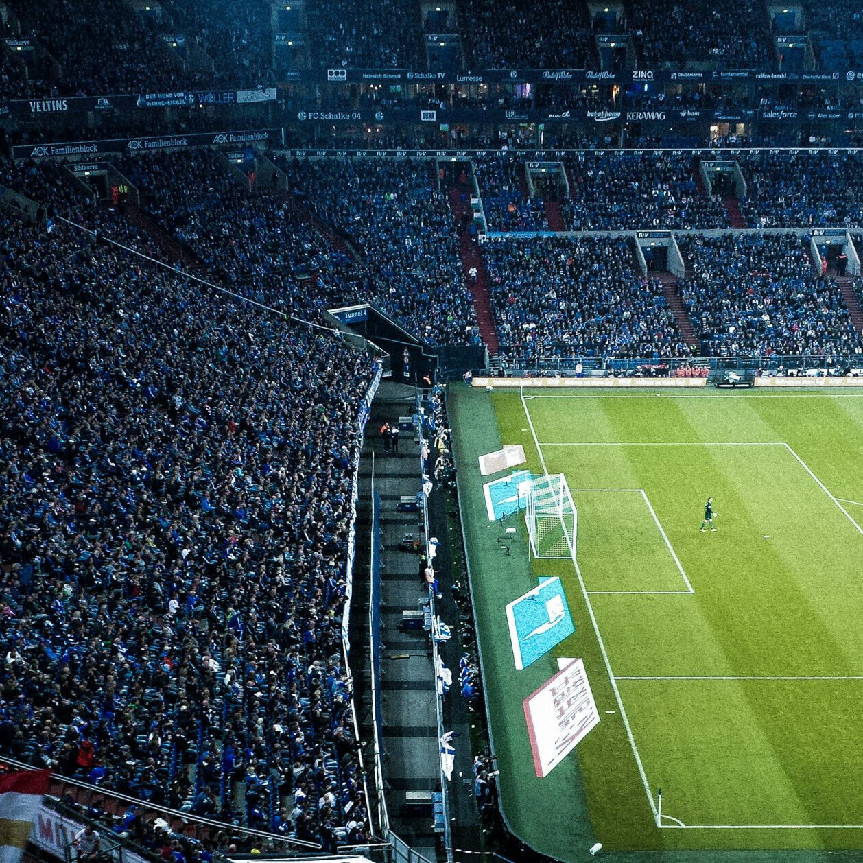 Ein vollbesetztes Fußballstadion mit Tausenden von Fans, die in den Tribünen sitzen. Das Spielfeld zeigt eine laufende Partie, bei der die Spieler in blauen und roten Trikots agieren. Im Vordergrund ist das Tor einer Mannschaft zu sehen. Das Stadion ist hell erleuchtet, und die Zuschauer tragen größtenteils blaue Fanbekleidung.